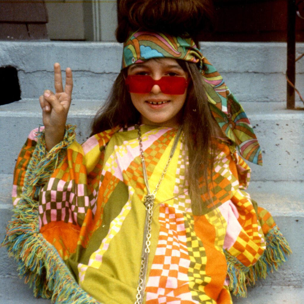 Gayle at 10 wearing rose-coloured glasses, a poncho, and a gold chain, flashing a peace sign